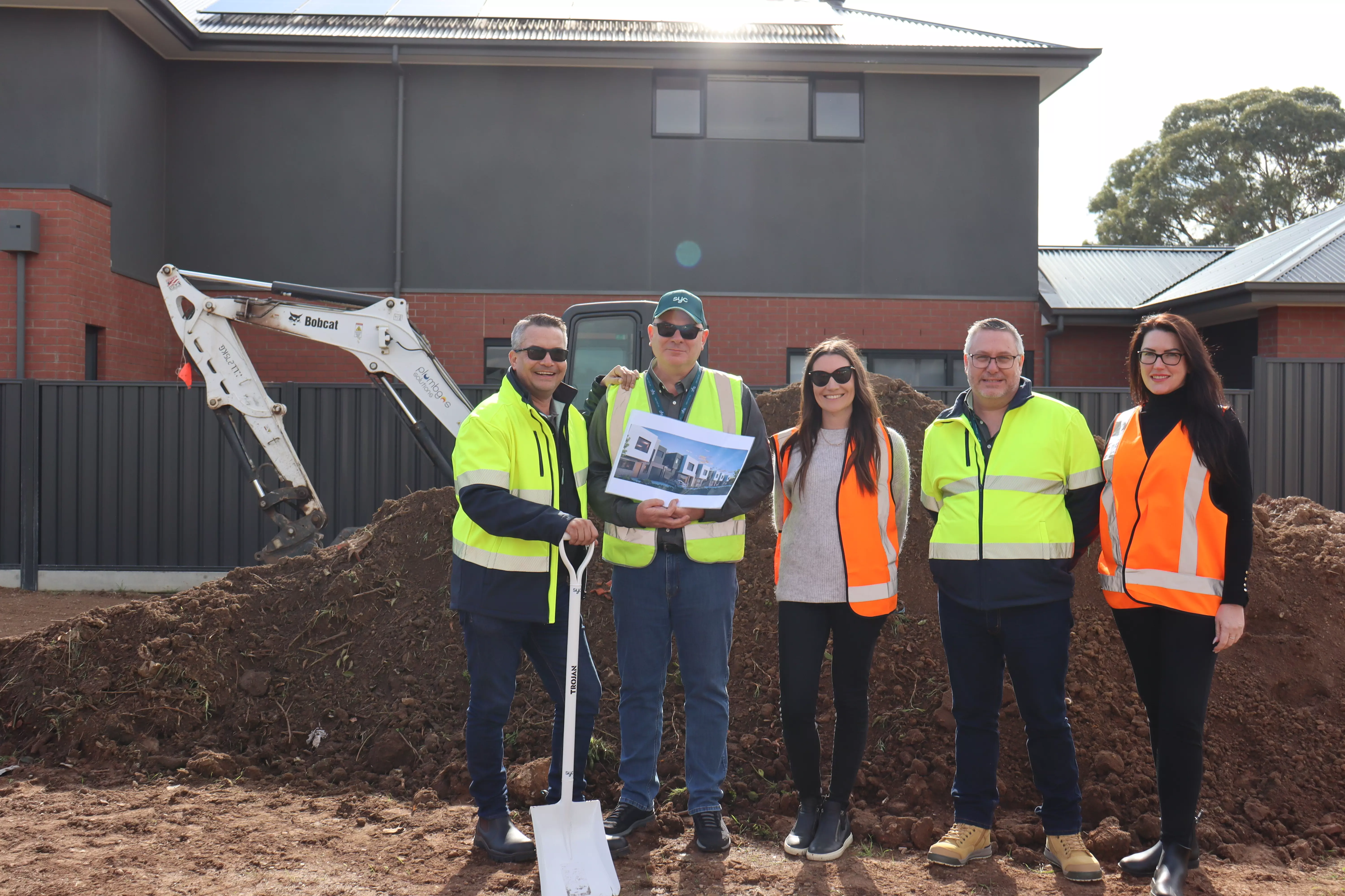 Five people wearing high-vis vests and safety gear standing on a construction site in front of a pile of dirt and an excavator. One person holds a printed image of a building design, and another holds a white shovel