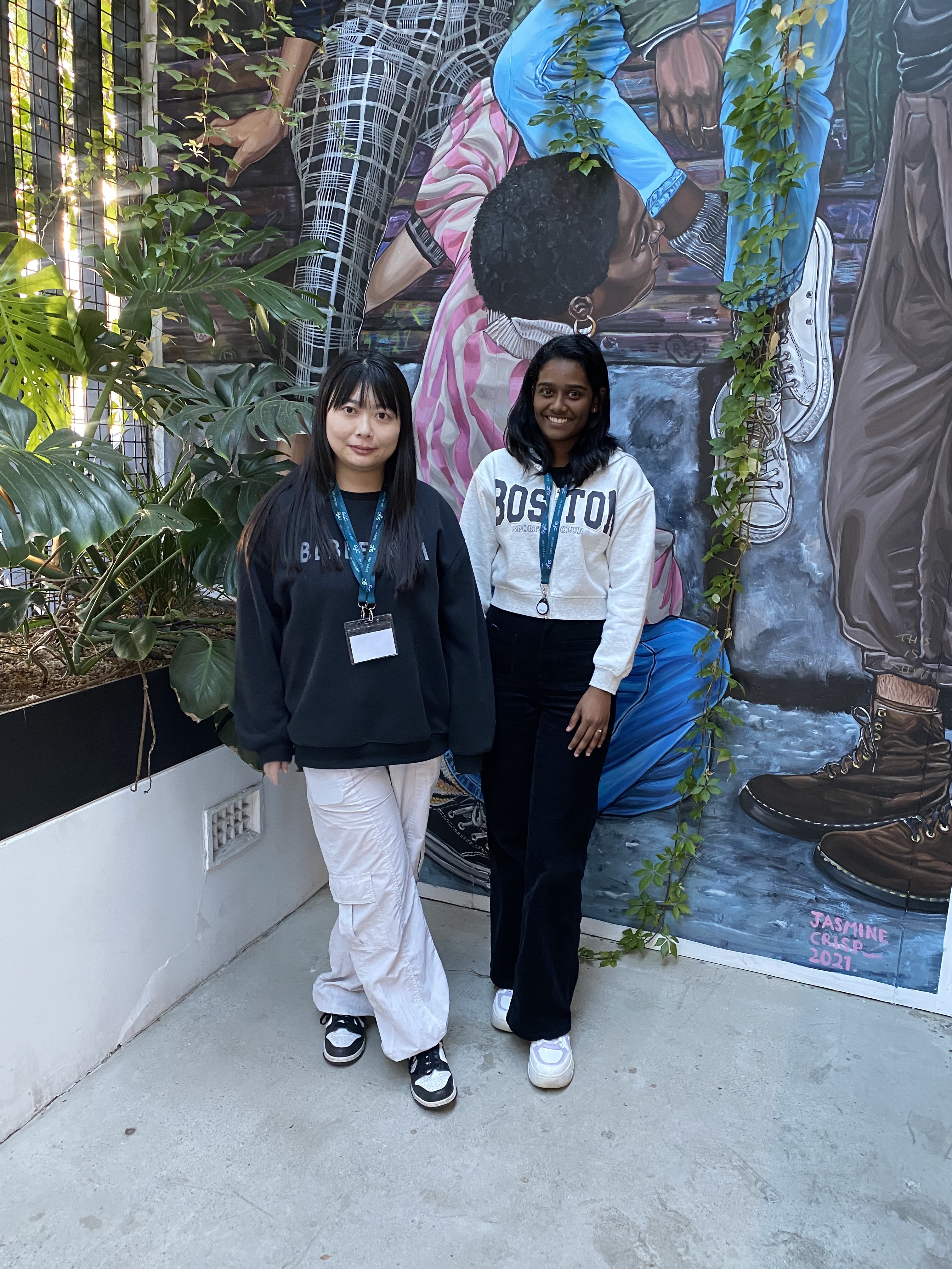 Two young women wearing lanyards standing in front of a colourful mural, surrounded by greenery.