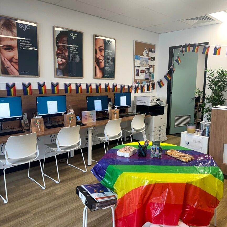 A decorated office space with computers along the wall, each with small rainbow flags above them. A central round table is covered with a rainbow pride flag and has snacks, stationery, and IDAHOBIT materials. Posters with smiling faces and SYC branding are displayed on the wall.