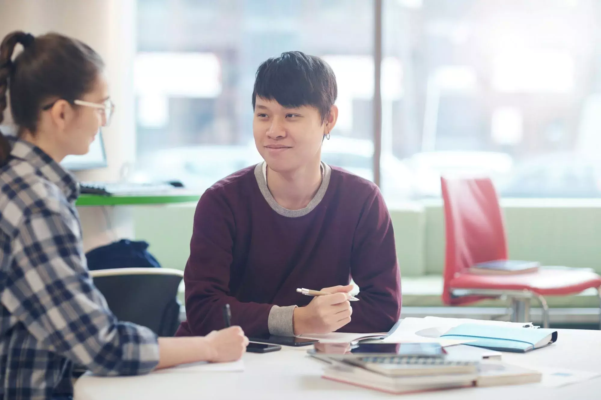 Two students sitting at a desk discussing work while one holds a pen.